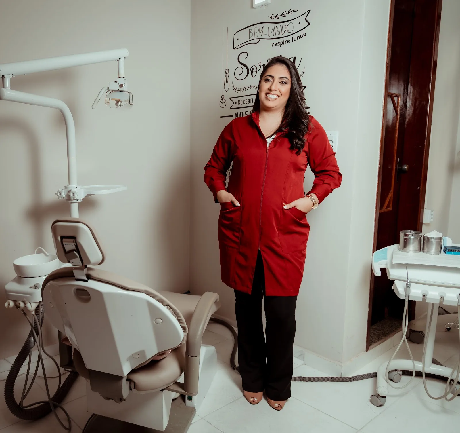 Dra. Bruna Geovana, de jaleco vinho, de pé no consultório ao lado da cadeira odontológica