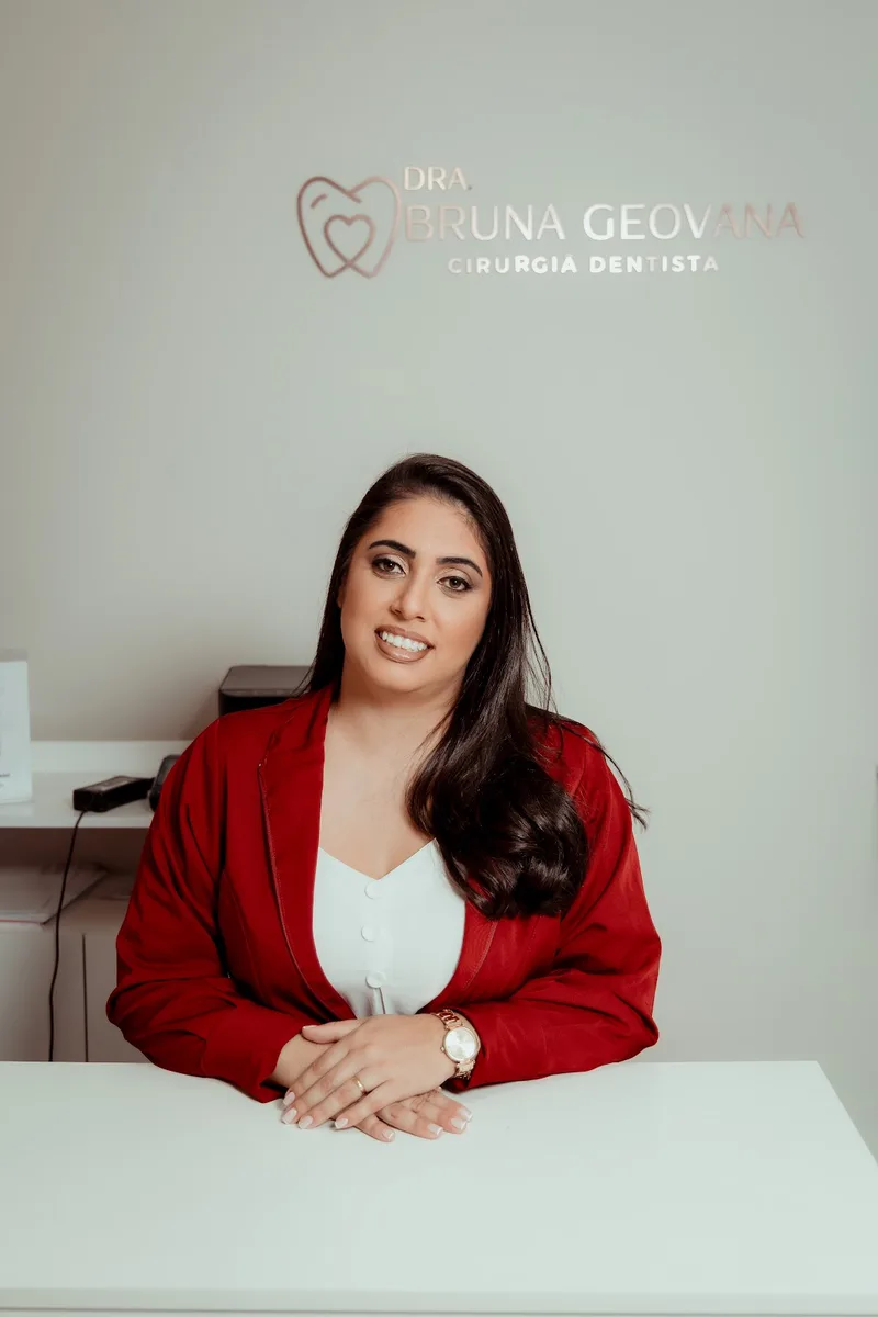 Dra. Bruna Geovana sentada à mesa do consultório, com o logotipo na parede ao fundo